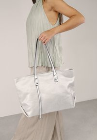 Bolso tote plateado con textura suave, que cuenta con dos correas largas y acentos negros contrastantes. Llevado por una persona con un atuendo claro.