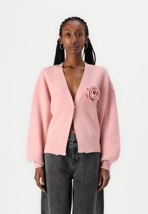 Rosa cardigan laget av myk stoff, med dyp V-hals, oversized ermer, ribbestrikkede kanter og en strikket rose detalj på forsiden.