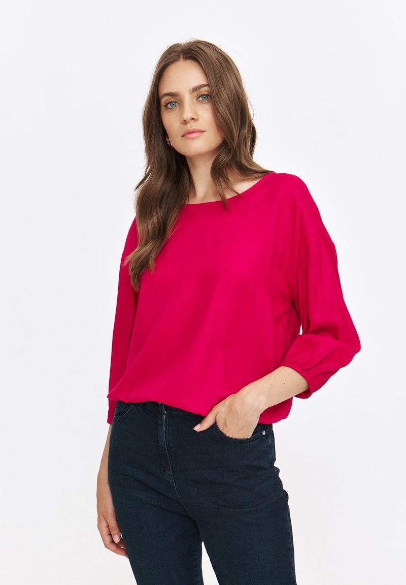 Fuksjowa bluzka o luźnym kroju, okrągłym dekolcie i marszczonych rękawach. Noszona z ciemnymi dżinsami, ma gładką teksturę.