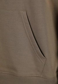 Tessuto di felpa marrone con tasca frontale. La texture è liscia e presenta cuciture a punto e un orlo a costine.