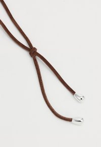 Corda in suede marrone con un nodo annodato, dotata di punte in metallo argento a entrambe le estremità. Texture liscia con un design piatto e flessibile.