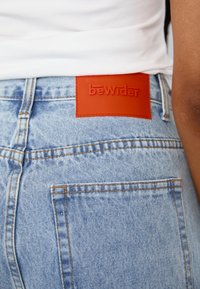 Lyseblå denimbukser med oransje søm og en rød lærpatch merket "beWider" i midjen; har baklommer.
