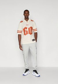 Κρεμ ποδοσφαιρική φανέλα με κοντά μανίκια και πορτοκαλί λεπτομέρειες, με μεγάλο "60" στο μπροστινό μέρος, συνδυασμένη με γκρι cargo φόρμες και ασπρόμαυρα αθλητικά παπούτσια.