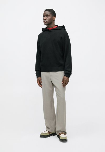 Tânăr îmbrăcat cu un hanorac negru, pantaloni bej largi și mocasini bicolori stând în fața unui fundal alb simplu.