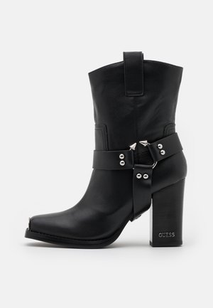 Stivale nero in pelle fino al polpaccio con tacco a blocco, borchie decorative in argento e fibbie, con la scritta "GUESS" in rilievo sul tallone.