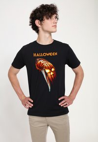 Černé bavlněné tričko s grafickým designem, na kterém je text "HALLOWEEN" nad stylizovanou rukou držící nožík a tvář dýně.