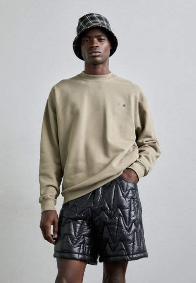 Olivgrüner Sweatshirt mit gerippten Bündchen, kombiniert mit schwarzen, gesteppten Shorts mit Zickzackmuster, Modell trägt einen karierten Bucket Hat.
