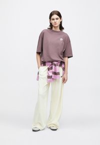 Mlada ženska stoji s rokama v žepih, oblečena v prenajit ovitek v barvi mauve, smetane široke hlače, superge in črtasto srajco, zavezano okoli pasu.