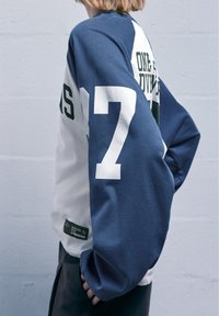 Cămașă cu mânecă lungă, cu blocuri de culoare albă și albastră, având numărul 7 mare, de culoare albă, și text verde. Material moale din bumbac, cu o croială lejeră.