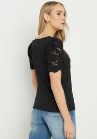 Schwarzes kurzärmliges Oberteil mit zarten Häkelspitzenärmeln mit floralen Mustern. Der Stoff ist glatt, und das Design ist tailliert.