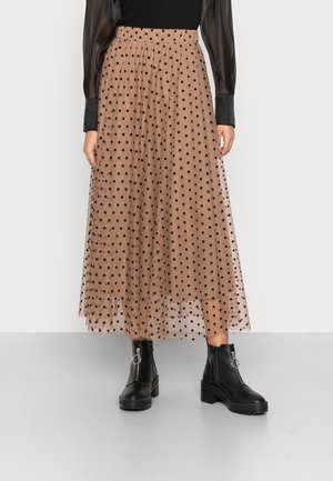 Kvinde iført en beige tyl-midi-nederdel med sorte polkaprikker, sort transparent langærmet top og sorte ankelstøvler med lynlåse.