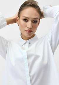 Biała koszula zapinana na guziki z klasycznym kołnierzem, długimi rękawami i subtelnym, srebrnym wzorem na całym materiale. Gładka faktura.