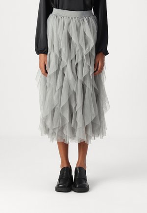 Personne portant une jupe midi en tulle gris superposée, un haut noir à manches longues et des chaussures noires sans lacets, debout devant un fond uni.