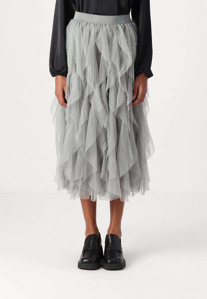 Personne portant une jupe midi en tulle gris superposée, un haut noir à manches longues et des chaussures noires sans lacets, debout devant un fond uni.