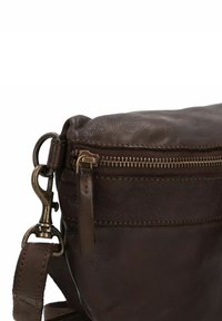 Angolo di una borsa in pelle marrone scuro con dettaglio di fibbia metallica, cerniera e tracolla su sfondo bianco.