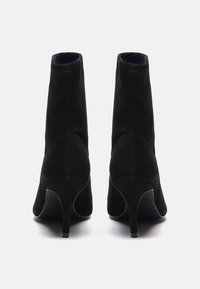 Bottes de cheville en suede noir avec un design ajusté, bout pointu et un talon haut élancé. La texture est lisse avec des détails de couture visibles.
