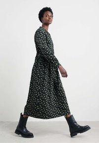 Bloemenprint, lange mouw jurk in donkergroen en beige, met een cinched taille en een vloeiende rok, gecombineerd met zwarte enkellaarsjes.