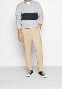 Grå tröja med dragkedja och blå rutigt panel, i kombination med beige cargobyxor med sidofickor och svarta låga sneakers.