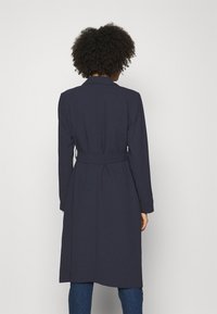 Cappotto blu navy con vita regolabile, maniche lunghe e colletto a risvolto. Tessuto liscio, lunghezza al ginocchio, con un design pulito e sartoriale.