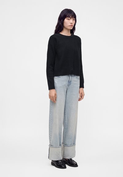 Femme debout portant un pull noir à manches longues, un jean large bleu clair avec de grands revers, et des mocassins noirs brillants sur fond blanc.