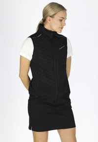 Schwarzes ärmelloses Vest mit gestepptem Muster, Reißverschluss und reflektierenden Akzenten. Über einem weißen Hemd getragen, mit schlankem Design.