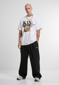 Balts t-krekls ar grafisko dizainu, kas iepazīstina ar vārdu "KIX" un dekoratīvu elementu, saderēts ar brīviem melniem sporta biksēm un sporta apaviem.