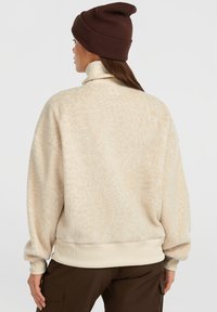 Vrouw in een beige fleece jas, crème coltrui, bruine beanie en bruine broek, van achteren weergegeven tegen een effen achtergrond.