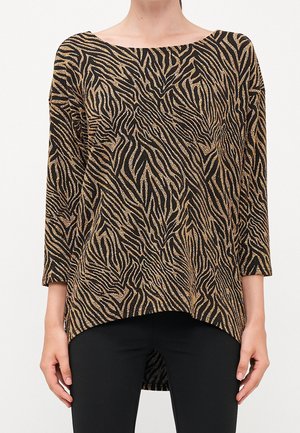 Nainen, joka pukeutunut pitkähihaiseen musta-kultaiseen eläinkuosisektopaan, jossa on venekaulus ja epäsymmetrinen helma, yhdistettynä mustiin housuihin.