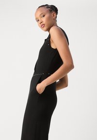 Robe noire sans manches à coupe slim, avec un décolleté arrondi, des poches latérales et des clous décoratifs sur les épaules et à la taille.