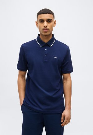 Άνδρας που φοράει μπλε μπλουζάκι πόλο με κοντά μανίκια και άσπρο περίγραμμα στο γιακά, στέκεται μπροστά σε απλό φωτεινό φόντο με τα χέρια στις τσέπες.