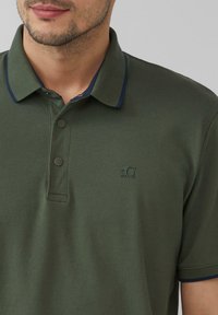 Groene polo met marineblauwe kraagaccenten, voorzien van twee knopen en een klein logo op de linkerkant van de borst. Gemaakt van een katoenmix.