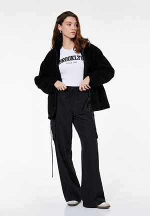 Kobieta ubrana w czarną kurtkę polarową, białą koszulkę z logo Brooklyn, luźne czarne spodnie i trampki, stojąca z rękami blisko talii.