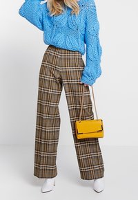 Jasně modrý, hrubě pletený svetr se vzory copánků; široké kalhoty s hnědo-béžovým kostkovaným vzorem; malá žlutá kabelka se zlatým řetízkovým popruhem.