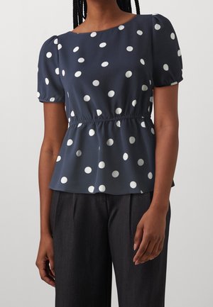 Femme portant un chemisier bleu marine à manches courtes avec des pois blancs, taille froncée, associé à un pantalon gris foncé, debout devant un fond uni.