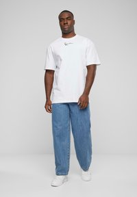 Biela bavlnená tričko s krátkymi rukávmi a jemným čiernym logom na hrudi, kombinované s voľnými modrými džínsy a bielymi teniskami.