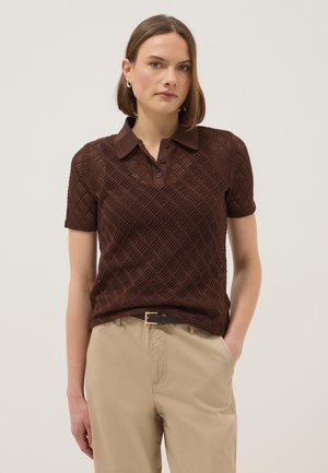 Femme portant un polo en maille à manches courtes de couleur marron foncé avec un pantalon beige, debout devant un fond clair uni.