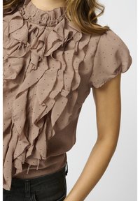 Blouse couleur blush en tissu léger avec des détails volantés et de petits pois noirs. Manches courtes avec un col haut.