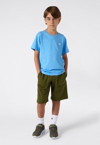 T-shirt en coton bleu clair avec un logo, associé à un short en polaire vert foncé avec de grandes poches, complété par des baskets grises et noires.