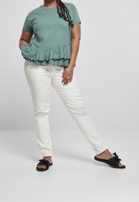Mujer vestida con una blusa corta de mangas rizadas en verde salvia, pantalones blancos y sandalias negras de slide con lazos, de pie contra un fondo claro y liso.