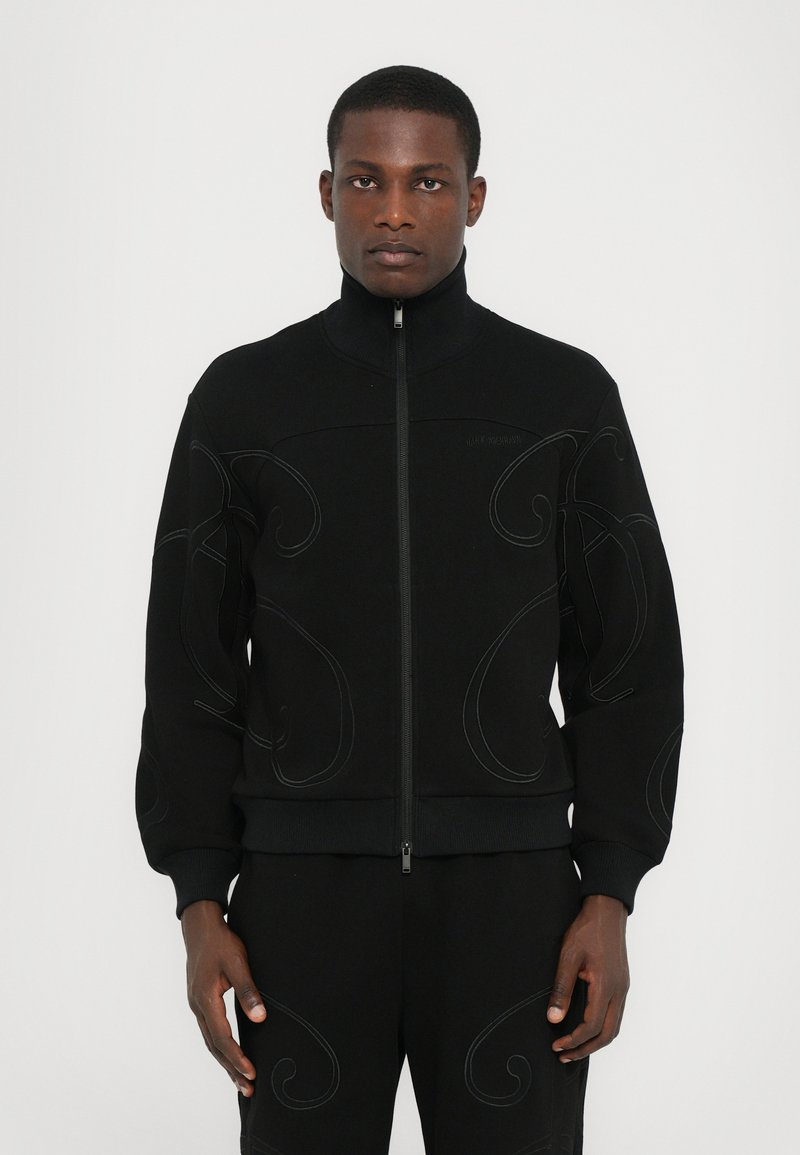 Sweatshirt zippé noir avec col haut, poignets côtelés et broderie décorative en fil ton sur ton légèrement contrasté.