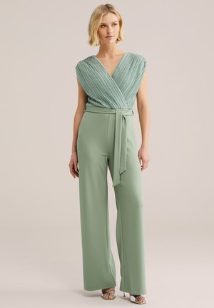 Kvinde iført ærmeløs lysegrøn plisseret omslagstop og matchende vidde bukser med bælte, stående mod en ensfarvet neutral baggrund.