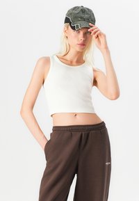 Hvit ribbestrikket crop tank topp kombinert med brune sweatpants med snøring. En grønn caps med hvite detaljer bæres, med buet skjerm og tekstilmateriale.