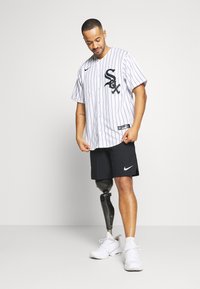 Férfi fehér baseball mez fekete csíkokkal és logóval, fekete Nike rövidnadrággal és fehér sportcipővel párosítva, bemutatva egy protéziskészüléket.