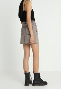 Minifalda de mezclilla en azul claro con dobladillo deshilachado, combinada con una camiseta sin mangas negra y botas negras con cordones. La vista posterior muestra detalles de la cintura y el ajuste.