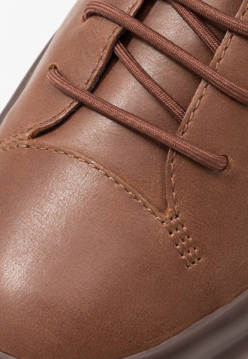 Zapato de cuero marrón con textura suave, que cuenta con seis agujeros para los cordones y cordones marrones. El detalle de costura a lo largo del área de los dedos añade interés al diseño.