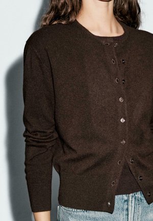 Personne portant un cardigan boutonné marron foncé sur un haut foncé et un jean bleu clair, avec de longs cheveux ondulés visibles.