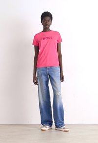 Pink cotton t-shirt with "BOSS" text, paired with light blue distressed wide-leg jeans. Model wears white and tan sneakers.