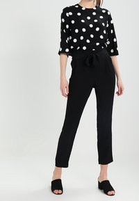 Chemisier noir à pois blancs avec manches trois-quarts, associé à un pantalon noir taille haute avec ceinture à nouer et jambes raccourcies.