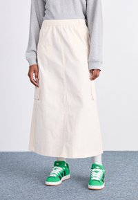 Beige midi szoknya, rugalmas derékkal, oldalsó zsebekkel és kontrasztos varrással, zöld sportcipővel és fehér zoknikkal párosítva.