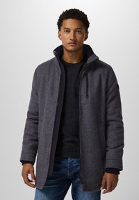 Cappotto in misto lana grigio con colletto alto, tasca con cerniera e polsini a costina, indossato sopra un maglione scuro e jeans blu.